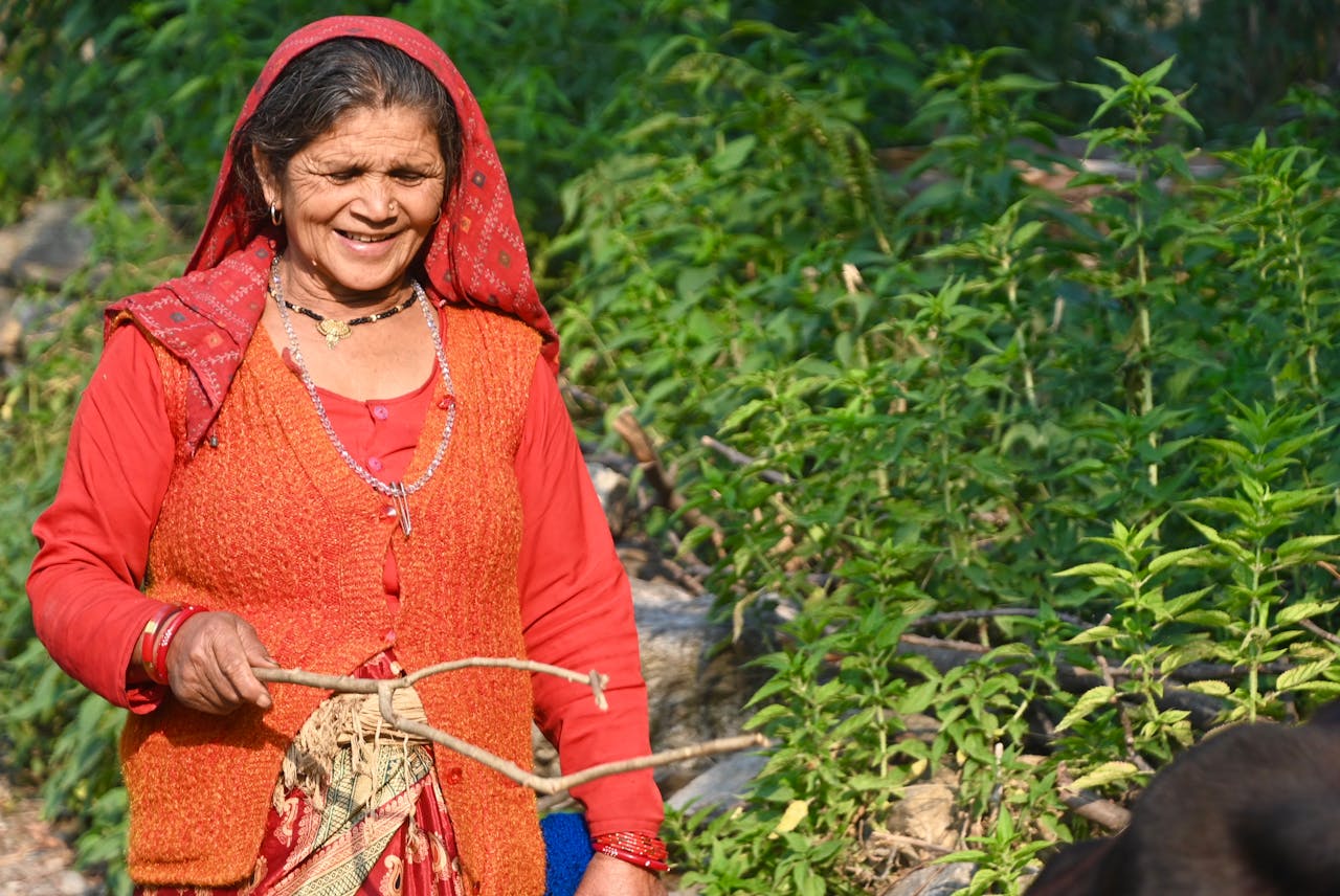 about-img Smiling elderly woman in traditional clothing holding a stick in a rural setting.