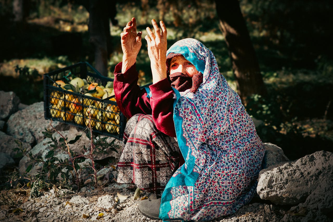Elderly woman wearing patterned shawl with fruit basket beside her outdoors.